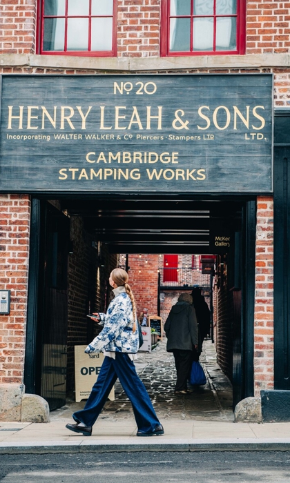 Closer view of the entrance to Leah’s Yard, showing the large wooden sign for ‘Henry Leah & Sons Ltd – Cambridge Stamping Works’ above the covered passageway. A cobbled walkway continues through to the courtyard, with brick façades and red-trimmed windows visible in the background.