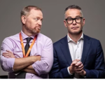 Studio portrait of two people seated side by side against a plain grey background. One wears a light purple shirt with an orange lanyard and sits with arms folded, looking towards the other person. The second wears a navy suit jacket, white shirt and dark-framed glasses, with hands clasped in front. The image has a clean, professional style with neutral lighting and no visible text.