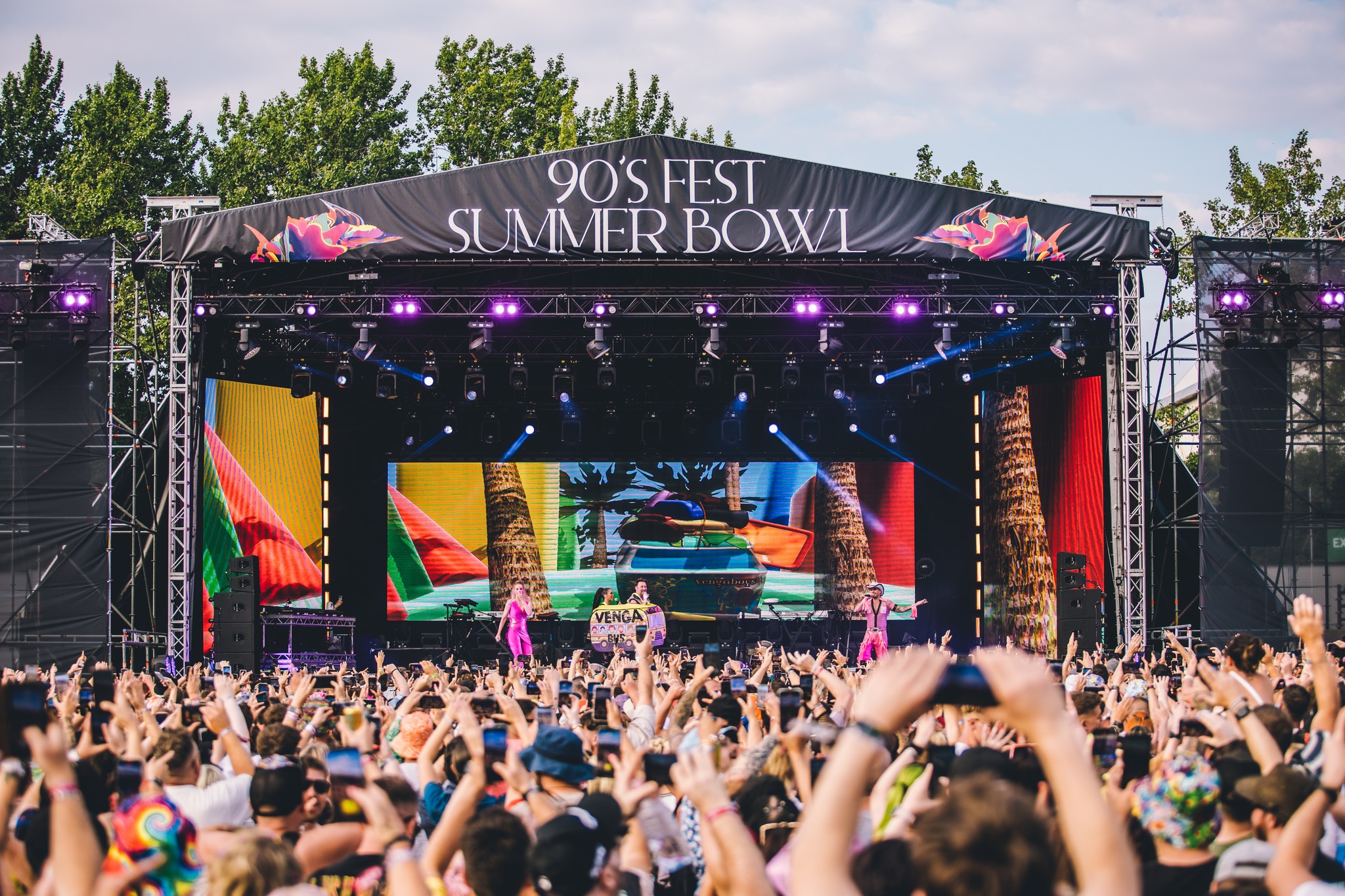 A large outdoor stage with a banner reading “90’s Fest Summer Bowl” features vibrant tropical graphics on big screens. Performers are on stage while a dense crowd in front raises hands and takes photos. Trees line the background under a partly cloudy sky.
