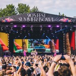 A large outdoor stage with a banner reading “90’s Fest Summer Bowl” features vibrant tropical graphics on big screens. Performers are on stage while a dense crowd in front raises hands and takes photos. Trees line the background under a partly cloudy sky.