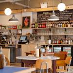 A bright, modern pizzeria interior with round tables, wooden chairs, hanging globe lights and shelves stocked with drinks behind the counter.