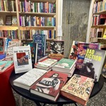 A round display table inside a bookshop showcasing a variety of new and featured books, arranged in stacks and upright stands. The table is surrounded by tall bookshelves filled with colourful books, creating a vibrant and busy bookstore interior.