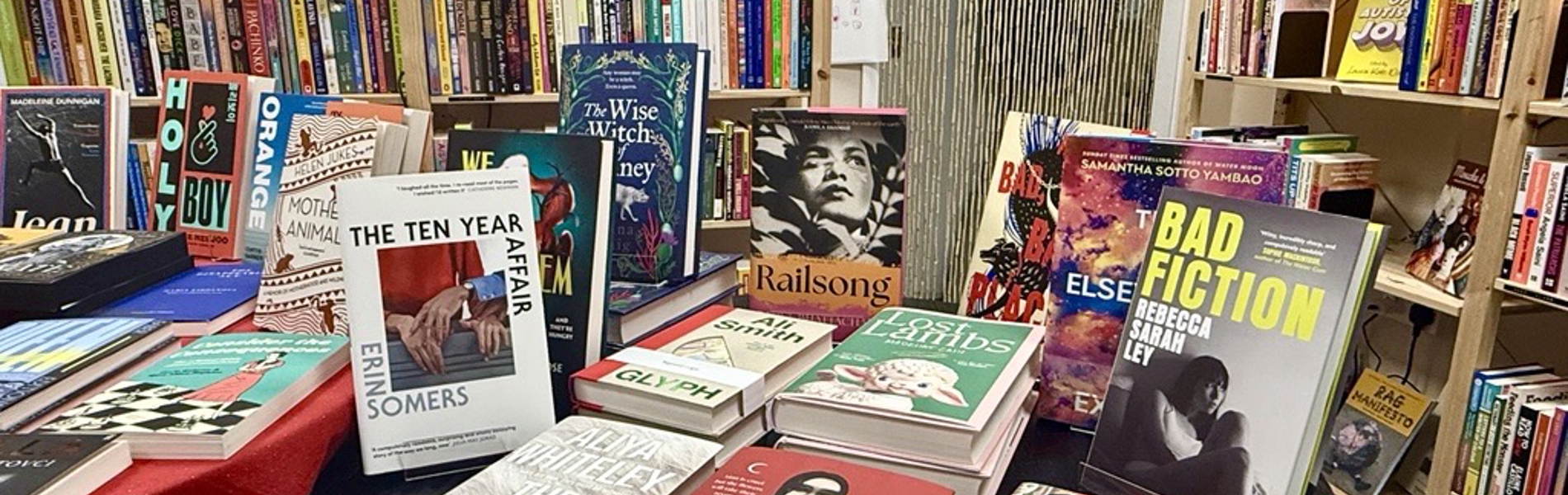 A round display table inside a bookshop showcasing a variety of new and featured books, arranged in stacks and upright stands. The table is surrounded by tall bookshelves filled with colourful books, creating a vibrant and busy bookstore interior.
