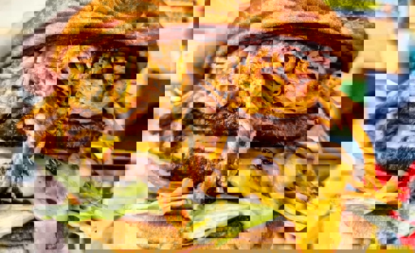 Close-up of a large burger held in one hand, featuring two beef patties layered with melted cheddar cheese, shredded lettuce, and a generous portion of pulled pork on top. The burger is in a toasted bun with a slightly cracked surface, and the background shows a wooden table with blurred food items and colorful packaging.