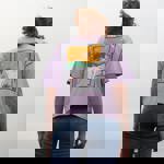 Person wearing a lavender oversized T-shirt with a bold graphic on the back featuring an abstract desert scene with a white goat, green mountains, orange sky, and geometric patterns. Paired with blue jeans, standing outdoors against a cloudy sky.