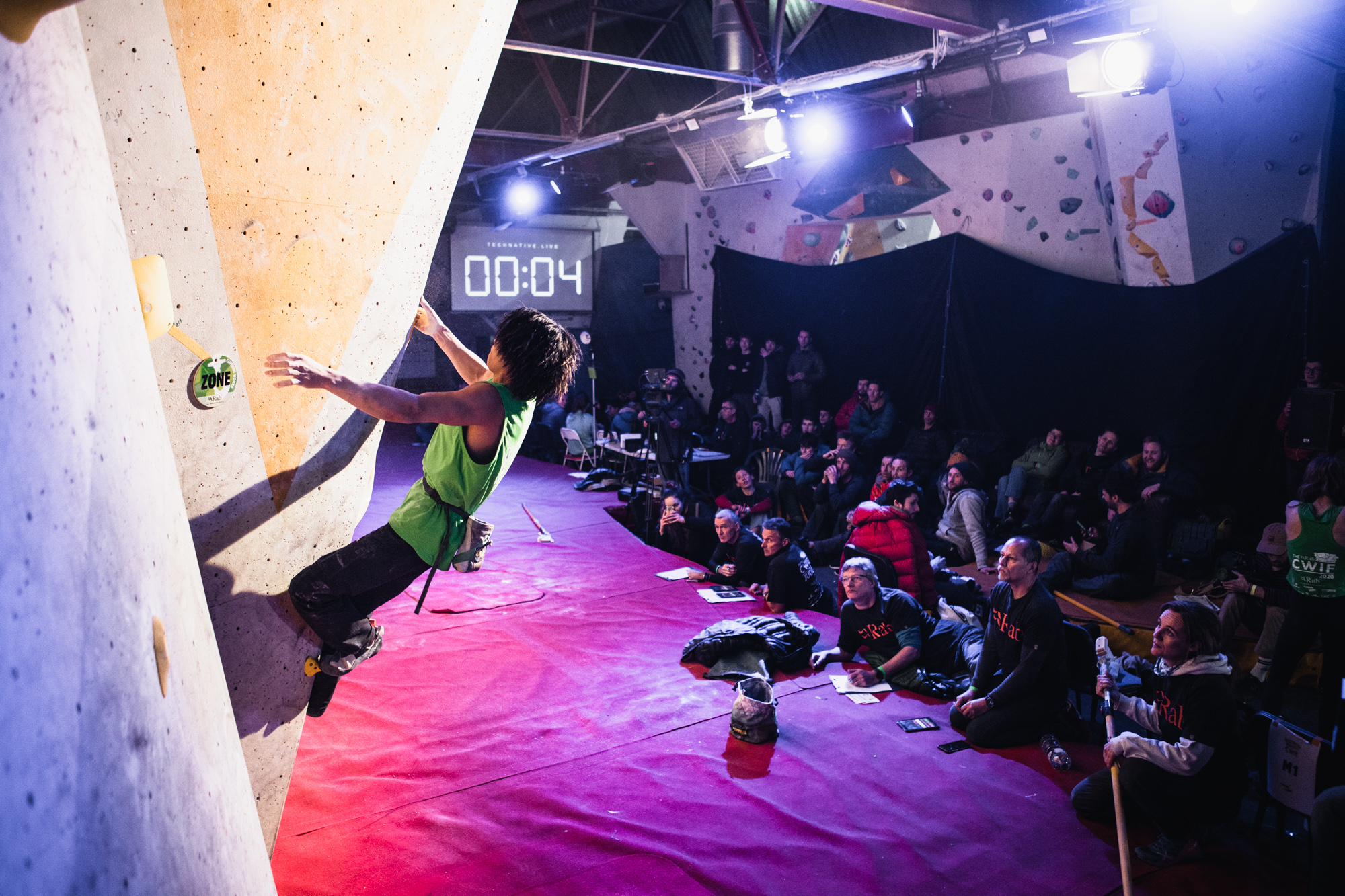 A man climbing on a wall. He has two feet and one hand on the wall, with his left hand reaching for the next hold. In the background, there is a digital timer displaying four seconds,and a crowd of people watching him.