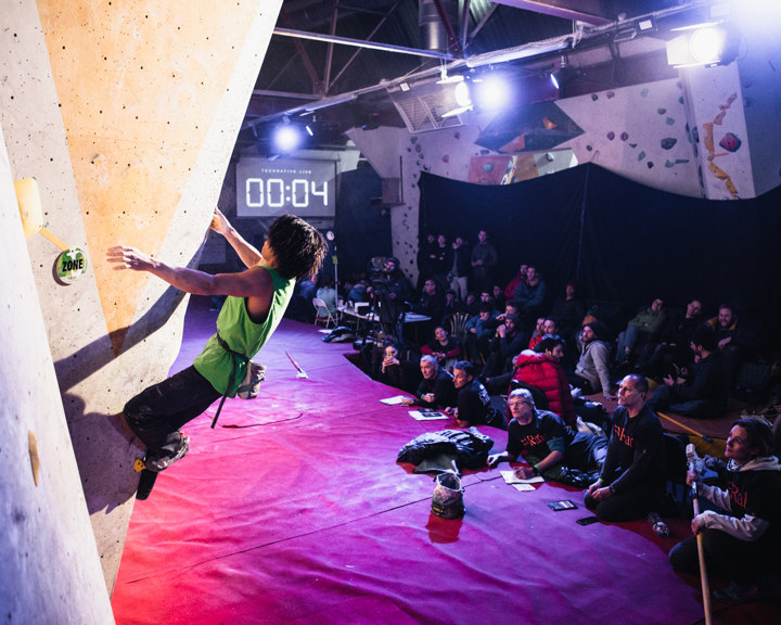 A man climbing on a wall. He has two feet and one hand on the wall, with his left hand reaching for the next hold. In the background, there is a digital timer displaying four seconds,and a crowd of people watching him.