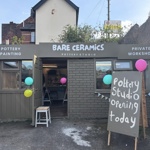 Exterior of Bare Ceramics pottery studio with open doors, colourful balloons, and a sign reading “Pottery Studio Opening Today,” showing the entrance and workshop space inside.