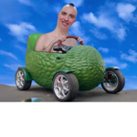 A surreal digital image showing a person seated in a small car shaped like a giant green avocado. The avocado vehicle has a textured exterior, shiny silver wheels and an open-top design with a steering wheel visible inside. The vehicle is parked on a sandy surface beneath a bright blue sky with scattered white clouds. The scene has a playful, computer-generated appearance.