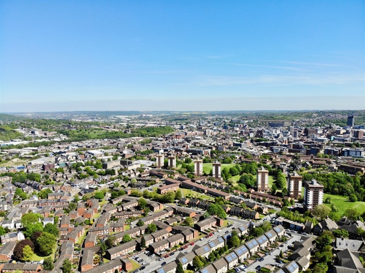 Aerial view of a cityscape on a clear day, featuring a mix of residential and commercial buildings. High-rise apartment blocks stand out among lower houses, with green spaces and trees interspersed throughout. The horizon stretches under a bright blue sky.