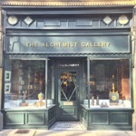 Exterior view of The Alchemist Gallery, an art gallery housed in a traditional shopfront with dark green painted woodwork and gold lettering above the entrance. Large display windows showcase ceramics, sculptures, framed artwork and decorative objects. The glass-panelled central door is framed by ornate stonework, and a doormat at the entrance reads “The Pharmacy”.
