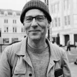A black and white head shot of a person standing in an urban street.
