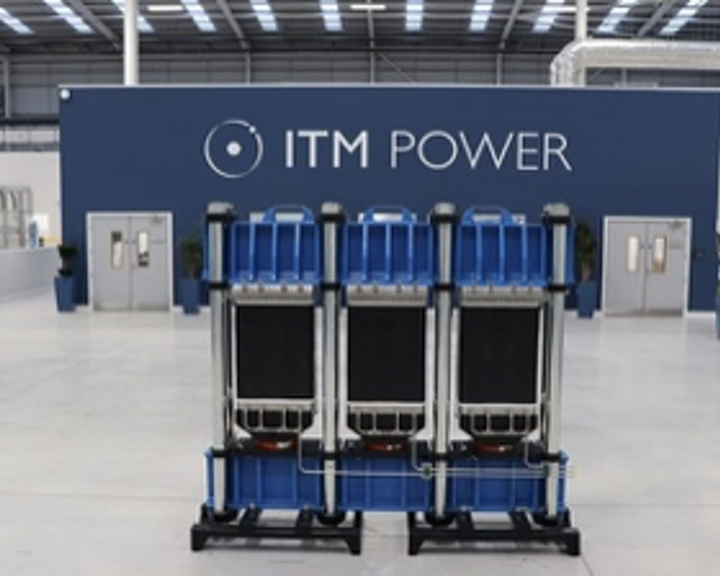 ITM Power: Industrial interior with a large blue and gray modular unit positioned in the center of a spacious factory floor. Behind it is a dark blue wall featuring the ITM Power logo in white. The space has high ceilings with metal beams, bright overhead lighting, and multiple doors and potted plants along the walls.
