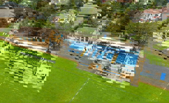 Aerial view of Hallam FC’s Sandygate stadium showing the main stand with blue seating and signage reading ‘Hallam F.C. Sandygate 1860.’ Below the stand are two dugouts, and to the left is a clubhouse building. The pitch is in the foreground, with trees and residential houses in the background.