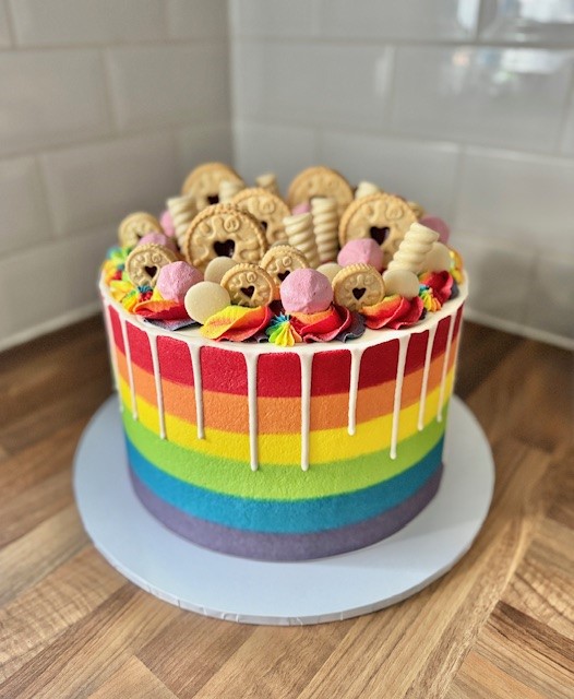 A vibrant vegan rainbow cake with horizontal stripes in red, orange, yellow, green, blue, and purple. The cake is topped with colorful buttercream swirls, pink marshmallows, and assorted biscuits, including jam-filled cookies and wafer rolls. White chocolate drip decorates the top edge.