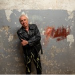 A person wearing a black leather jacket and patterned trousers stands with folded arms against a weathered grey wall with peeling paint and patches of exposed plaster. A red paint mark appears on the wall to one side. The portrait is taken indoors with soft lighting, highlighting the textured surface of the wall and creating a striking, urban backdrop.