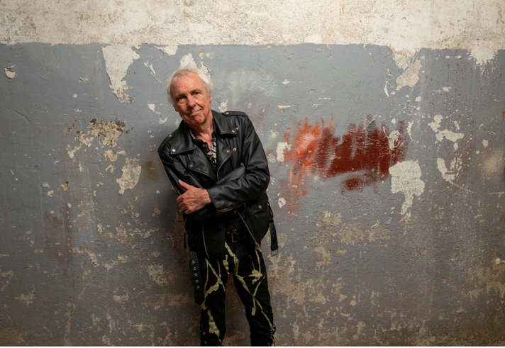 A person wearing a black leather jacket and patterned trousers stands with folded arms against a weathered grey wall with peeling paint and patches of exposed plaster. A red paint mark appears on the wall to one side. The portrait is taken indoors with soft lighting, highlighting the textured surface of the wall and creating a striking, urban backdrop.