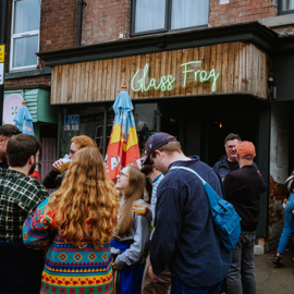 Group of people gathered outside a venue with a wooden facade and a neon sign reading ‘Glass Frog’. The entrance is flanked by colorful umbrellas, and the setting appears to be a lively street with adjacent shops and signage.