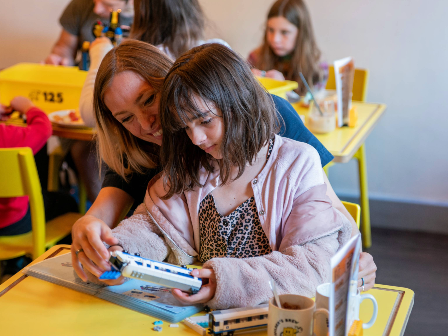 A person and a child are sat at a wooden table in a cafe, building a Lego© train together.