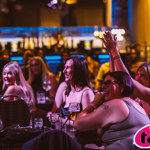 Audience members seated at tables in a dimly lit entertainment venue watch a live performance. In the foreground, several people sit with drinks, including beer glasses and cans, while one person raises a hand in the air. Coloured stage lighting in pink, blue and amber tones illuminates the scene, with a blurred bar and crowd visible in the background. A pink and black Tasty logo appears in the bottom-right corner.