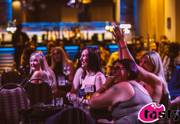 Audience members seated at tables in a dimly lit entertainment venue watch a live performance. In the foreground, several people sit with drinks, including beer glasses and cans, while one person raises a hand in the air. Coloured stage lighting in pink, blue and amber tones illuminates the scene, with a blurred bar and crowd visible in the background. A pink and black Tasty logo appears in the bottom-right corner.