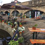 Outdoor seating area in front of a stone building with large archways. The space features wooden picnic-style tables with orange tops & green metal legs, some under large branded umbrellas. Colourful potted flowers line the edge of a canal in the foreground, adding vibrant reds, whites, and purples. Behind the seating area, the building has signage and posters visible through the arches, suggesting a café or bar setting. The ground is paved with cobblestones, and the overall scene has a relaxed atmosphere.