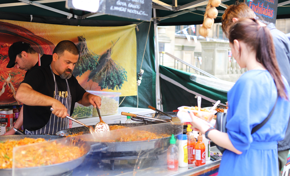 A street food trader stirs a large dish of paella as people watch 