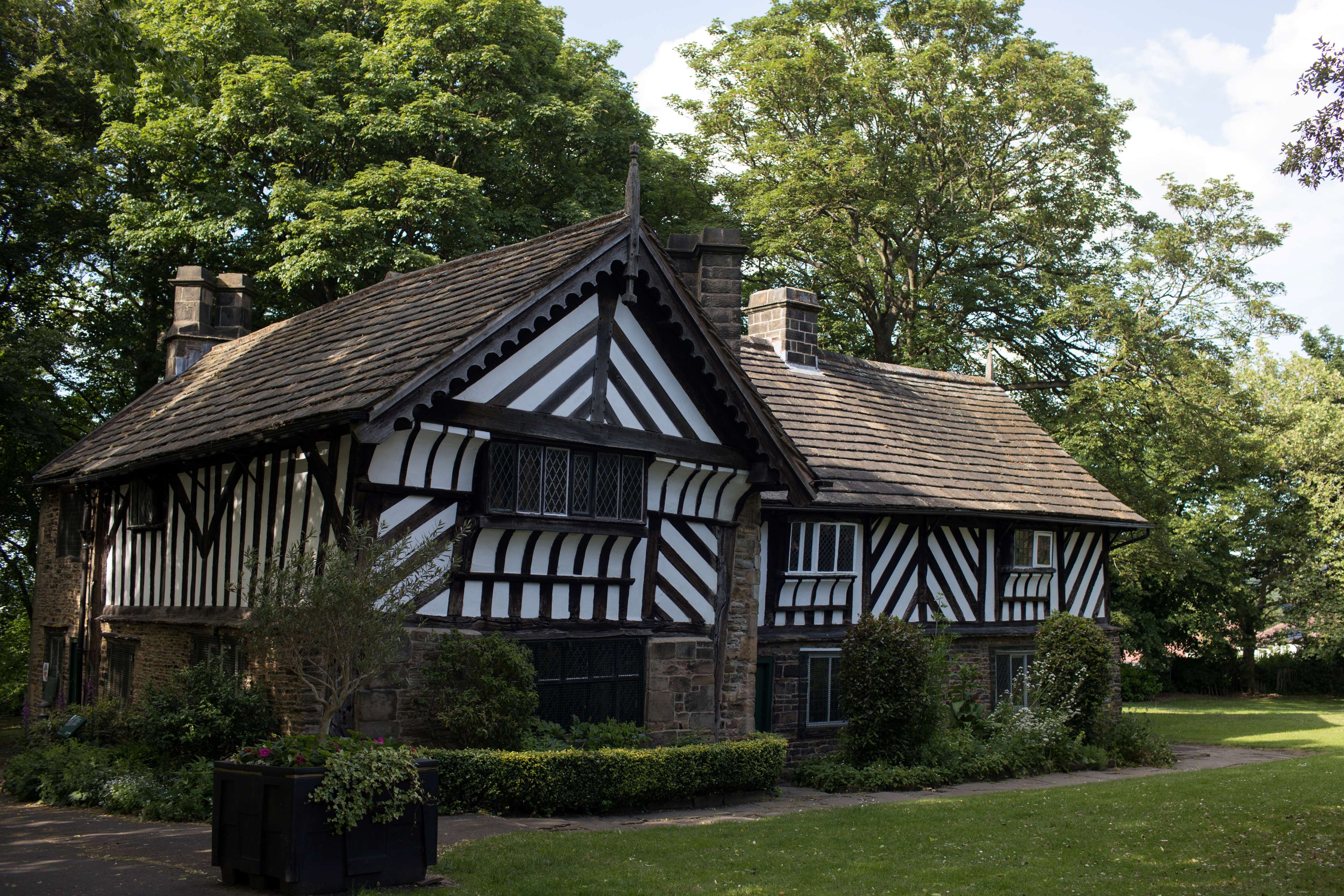 A historic black-and-white timber-framed building with a steep tiled roof sits in a green park surrounded by tall trees. The structure features decorative diagonal beams, small leaded windows, and stone walls at the base. A manicured lawn and shrubs frame the front, with a paved path leading toward the building under a bright, partly cloudy sky.
