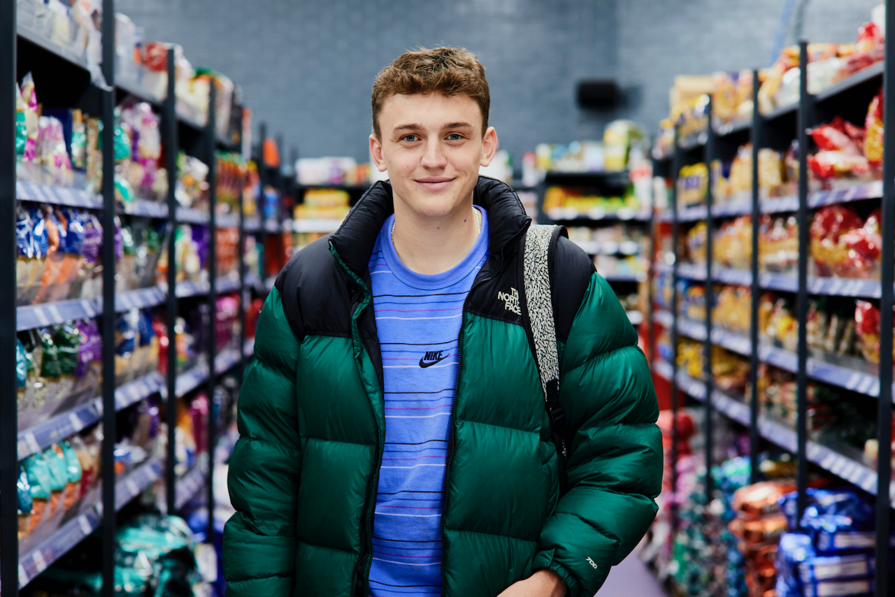 A person wearing a green puffer jacket over a blue striped Nike shirt is standing in the aisle of a grocery store. Shelves on both sides are stocked with colorful packaged snacks and food items, and the background shows more shelves filled with products under bright indoor lighting.