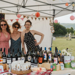 Three people in a marquee at an outdoor event. In front of them is a table filled with bottles of wine.
