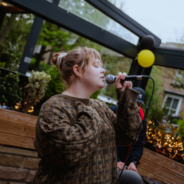 Person holding a microphone and performing outdoors in a covered patio area decorated with plants, string lights, and black and yellow balloons. Wooden planters and greenery surround the space, with buildings visible in the background