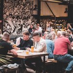 Crowds of people sat on benches drinking beer indoors, with a black and white graffiti wall in the background