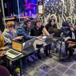 Eight people, all wearing headphones, are sitting round a large table working on four laptops.