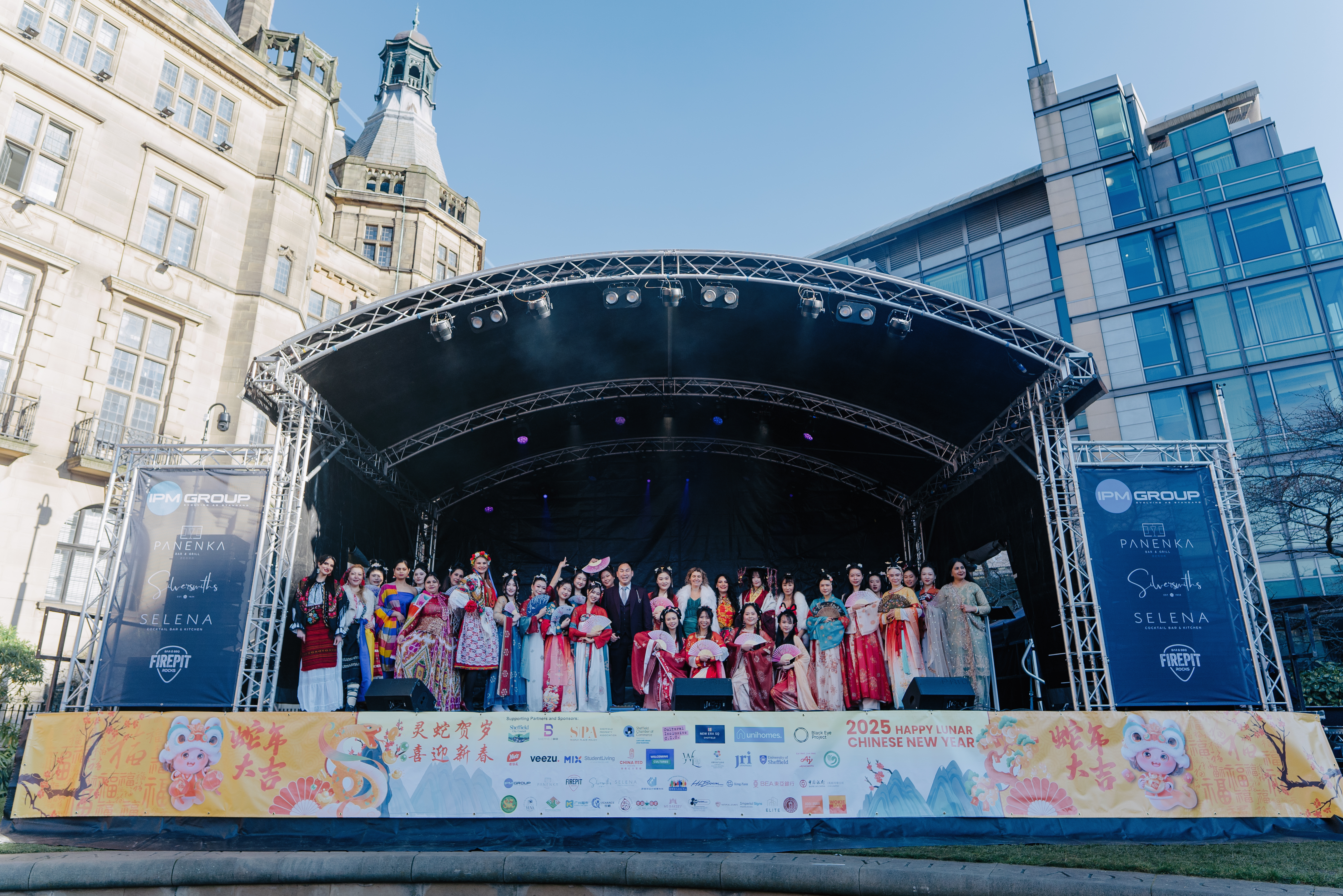 A large outdoor stage with a group of performers dressed in colorful traditional Chinese attire, standing in a line. The stage is decorated with banners and sponsor logos, and the backdrop shows historic and modern buildings under a clear blue sky.