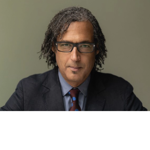 tudio portrait of a person wearing a dark suit, blue shirt and patterned tie, seated against a plain grey background. The person has shoulder-length textured hair and rectangular glasses, looking directly towards the camera. Soft, even lighting highlights the face, creating a professional and formal appearance.