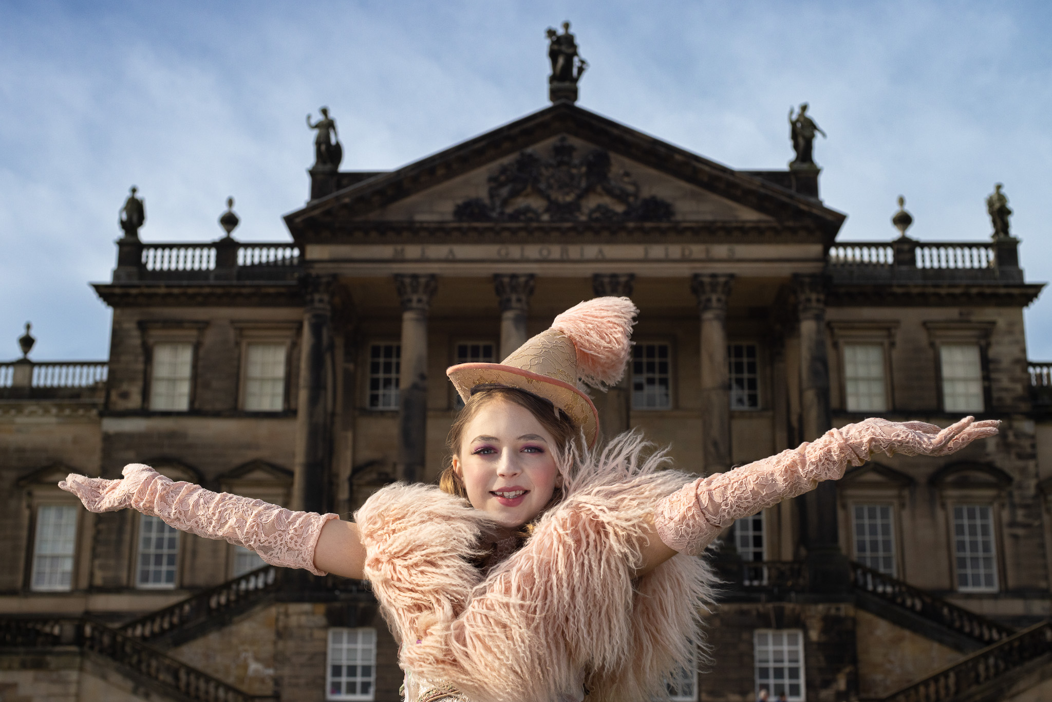 A performer in an elaborate, feathered costume stands with arms outstretched in front of a grand historic building with stone columns, statues and ornate architectural details. The outfit includes long lace gloves and a decorative hat, creating a dramatic, theatrical pose against the backdrop of the building.