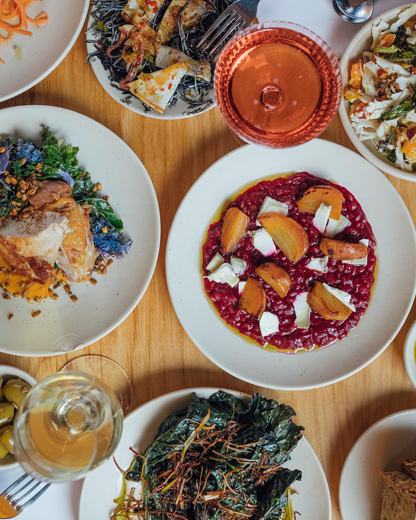 Top-down view of a wooden table set with several plates of colorful dishes, including a vibrant beetroot risotto topped with roasted golden beets and cheese, a plate of leafy greens with crispy garnish, and another with roasted meat and vegetables. Two glasses of wine—one white and one rosé—are also visible, along with small bowls of sides and garnishes.