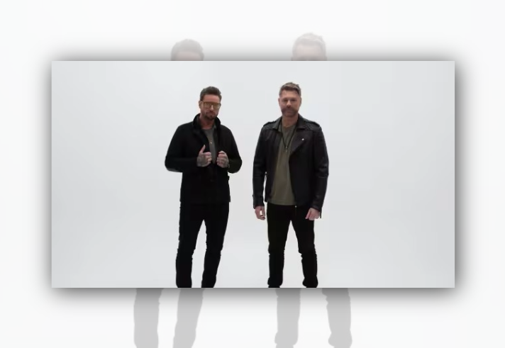 Two people stand side by side against a plain light-grey background. Both wear dark casual clothing, including jackets, slim-fit trousers and boots. One stands with hands raised near the jacket collar, while the other stands with arms relaxed by the sides. The minimalist studio-style image has no visible props, text or background details.