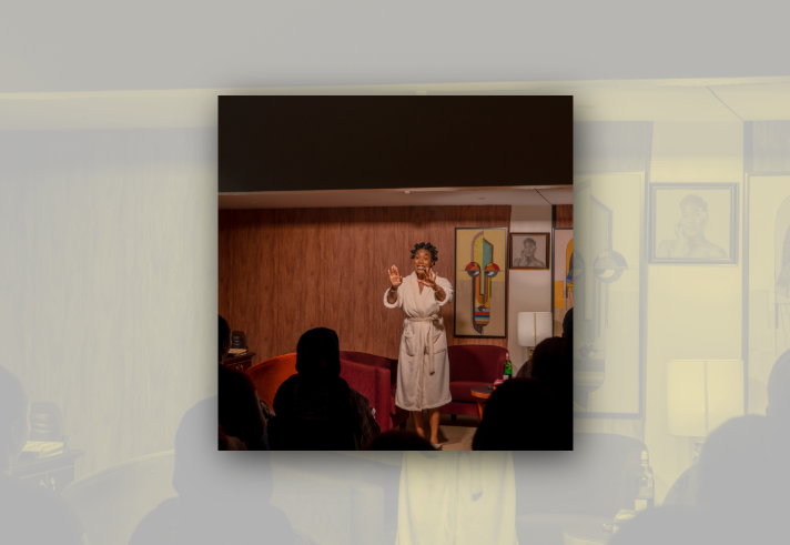 A performer stands barefoot in a warmly lit room set up like a living space, wearing a light-coloured dressing gown and addressing a seated audience. Red armchairs, a side table with a bottle, a floor lamp and framed artwork decorate the wood-panelled backdrop. Audience members appear in silhouette in the foreground, watching the intimate live performance.