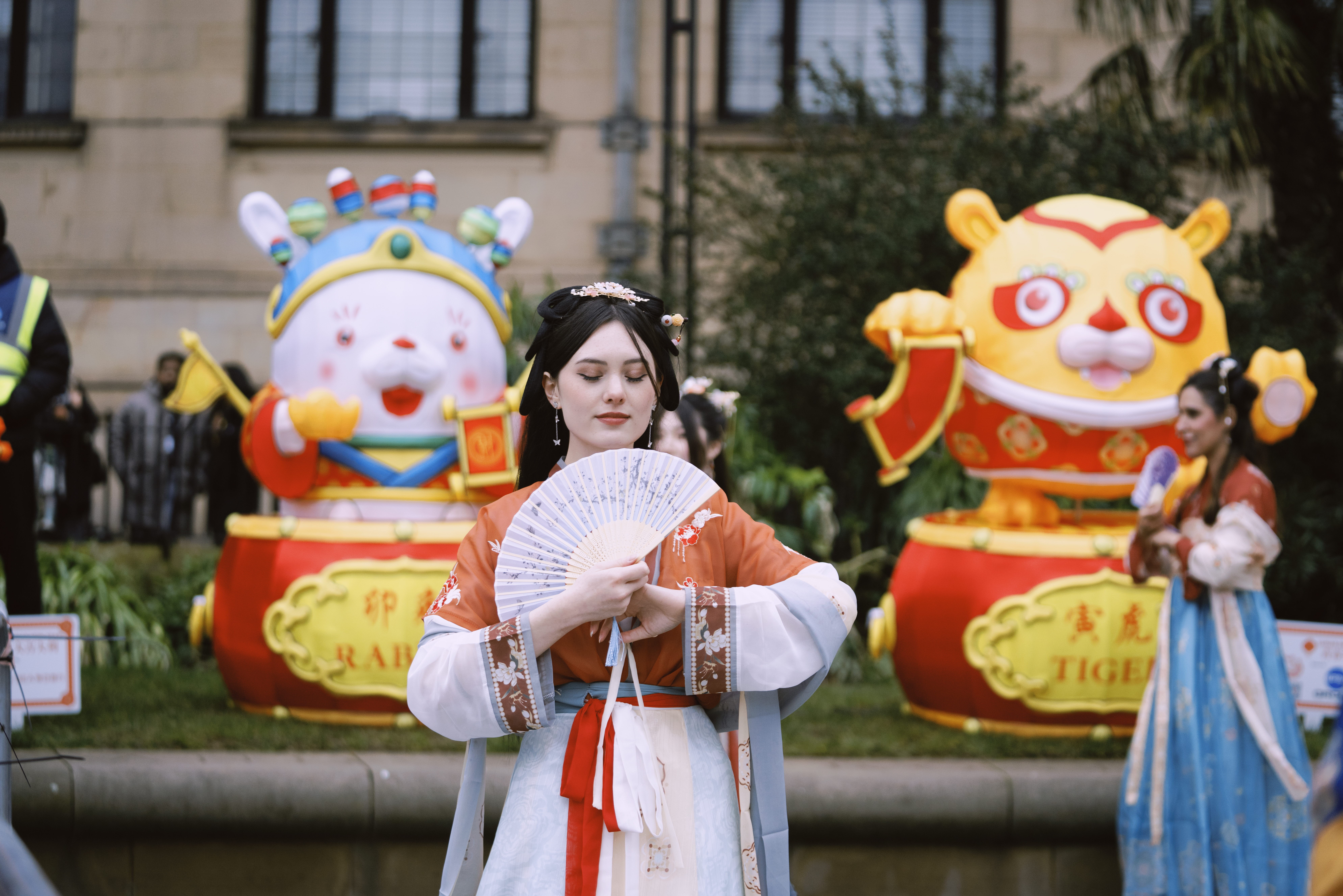 A person in traditional Chinese clothing holding an ornate folding fan, standing in front of large decorative figures representing the zodiac animals Rabbit and Tiger. The figures are colorful and festive, placed in an outdoor setting.