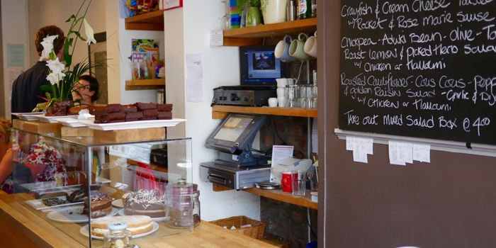 A counter in a cafe, with a glass case filled with cakes. Behind the counter is a menu written on a large blackboard.
