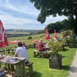 People sitting at wooden picnic tables in The Old Horns Inn beer garden on a sunny day, surrounded by red parasols, green lawns and sweeping views of the rural landscape.