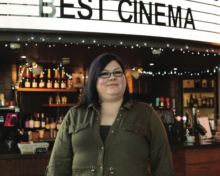 Person standing in front of a bar counter inside a cinema, with a large illuminated sign overhead reading ‘THE LIGHT SOCIAL SHEFFIELD AWARD BEST CINEMA.’ Shelves behind the counter display bottles and glasses, and string lights add decoration.