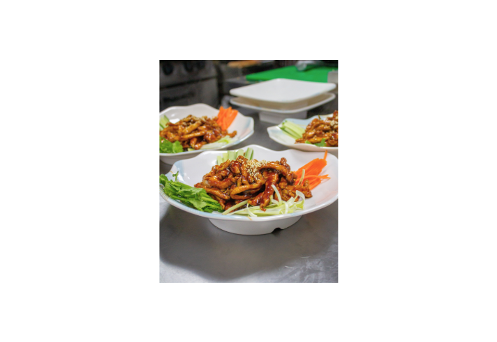 Three plated servings of a glossy brown stir-fried dish are arranged on a stainless-steel kitchen counter. The foreground plate is garnished with fresh coriander, shredded cucumber, carrot and sesame seeds, while two similar plates sit slightly out of focus in the background. The image highlights the presentation and preparation of the meal in a restaurant kitchen.