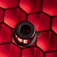 Close-up of a small cylindrical electronic component mounted on a surface with a red honeycomb pattern. The component has a metallic finish and a central lens-like opening, reflecting the red light from the background.