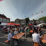 A busy pub beer garden filled with people seated at long wooden benches under colourful bunting and umbrellas, with houses and buildings visible around the courtyard.