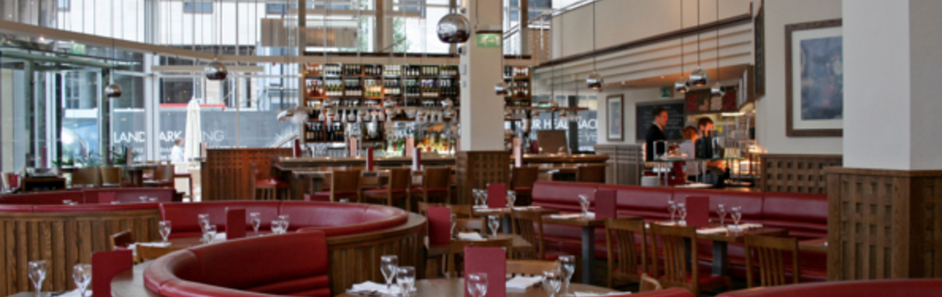 The interior at Piccolino. Bright, modern restaurant interior with large windows, high ceilings, and hanging pendant lights. The space features curved wooden booths with red leather seating and neatly set tables with glassware and menus. A long bar at the back is stocked with bottles, and the overall design combines warm wood tones with contemporary elements.