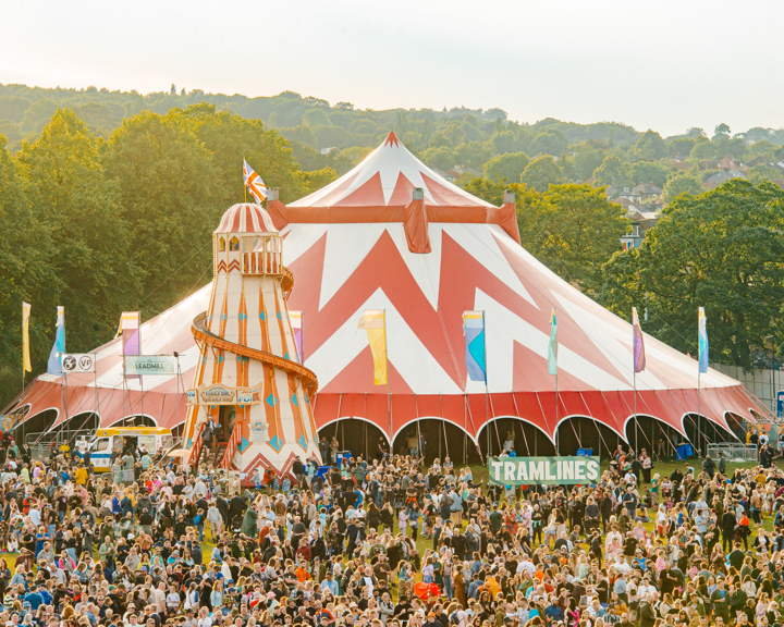 A huge red and white circus tent sits in the middle of Hillsborough Park. It is surrounded by huge crowds of people. There is a large banner visible that reads 'Tramlines'.