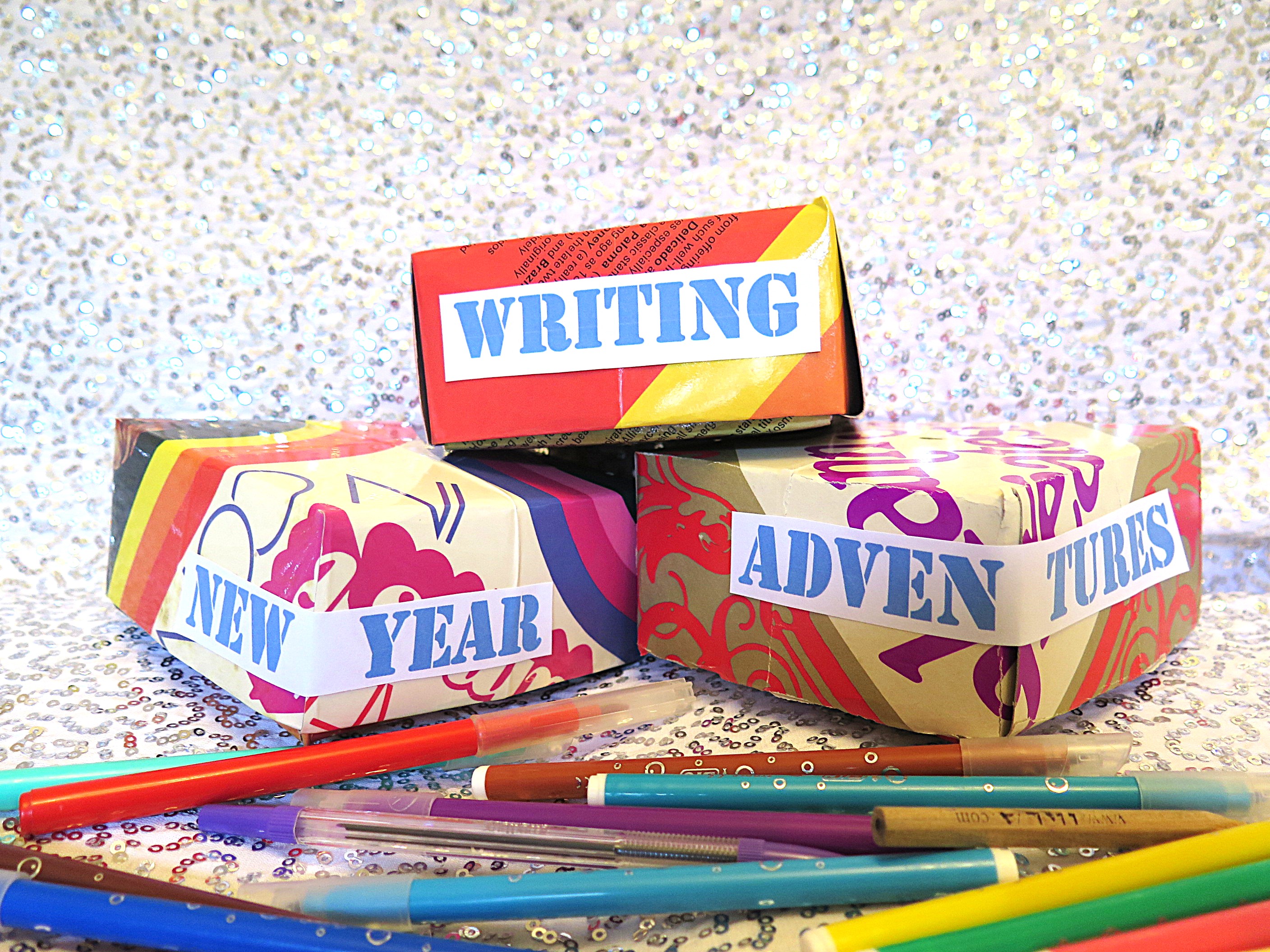 Three brightly coloured, patterned boxes sit against a sparkling silver backdrop. Each box carries part of the phrase “Writing New Year Adventures” in bold blue letters. A scattering of colourful pens lies in the foreground, adding to the creative, celebratory feel of the scene.