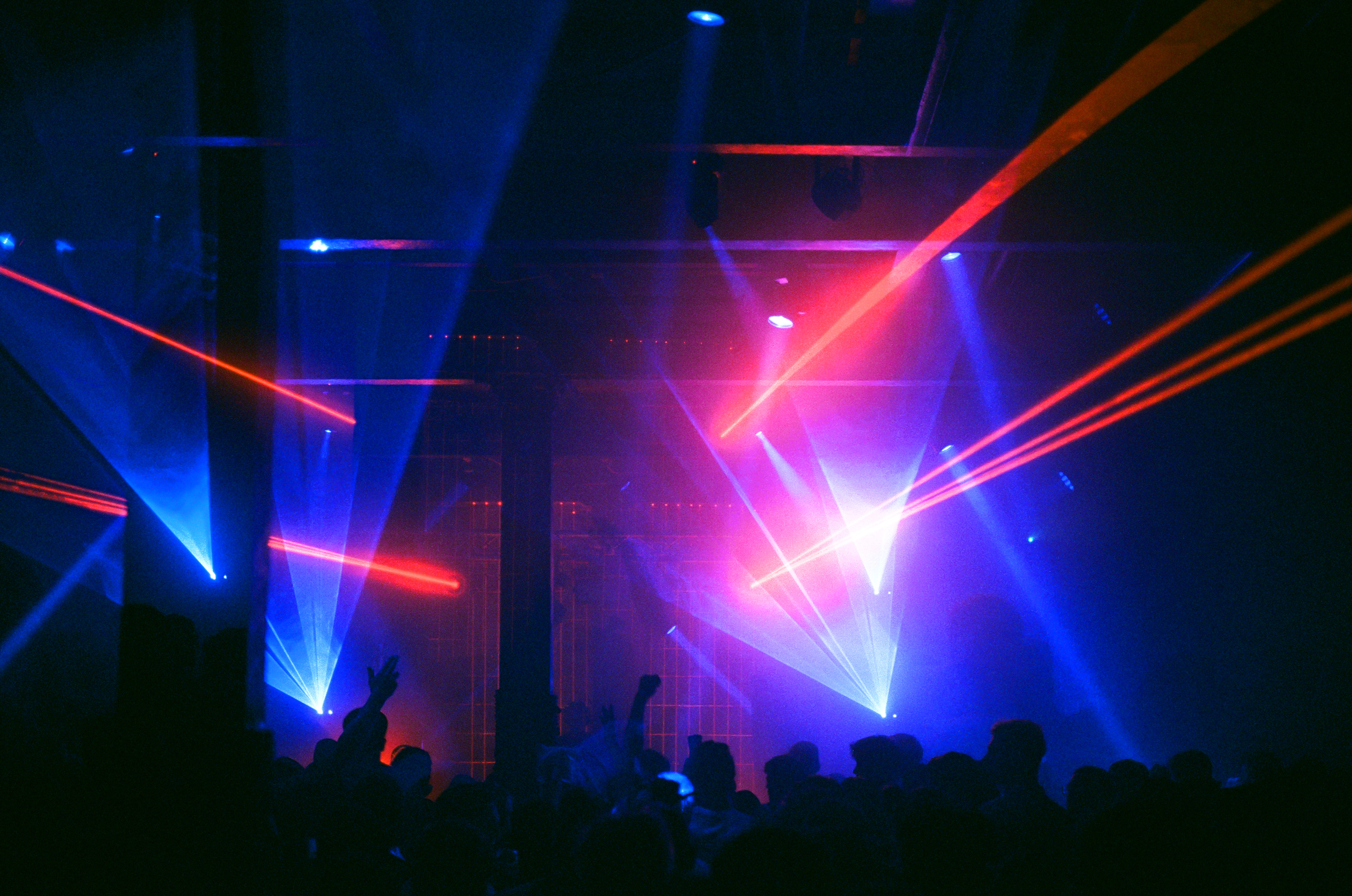 A crowded nightclub or music venue with vibrant laser lights in blue and red cutting across the dark space. Silhouettes of people are visible in the foreground, some with raised arms, creating a lively atmosphere. The beams of light converge toward the center stage area, highlighting the industrial-style ceiling and pillars. The overall scene conveys an energetic party or concert setting with dramatic lighting effects.